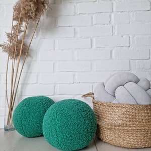 May include: Two round green fuzzy poufs or ottomans, a woven wicker basket with a gray knotted pillow inside, and a dried pampas grass arrangement in a clear vase on a white brick wall background.