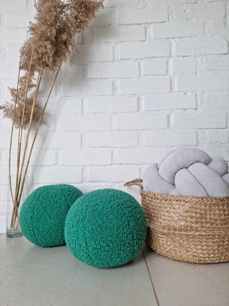 May include: Two round green fuzzy poufs or ottomans, a woven wicker basket with a gray knotted pillow inside, and a dried pampas grass arrangement in a clear vase on a white brick wall background.