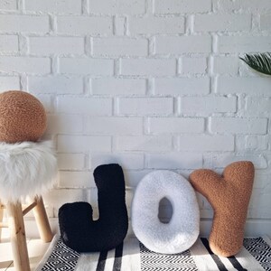 May include: Three large plush letters spelling out the word "JOY" in black, white, and brown. The letters are sitting on a black and white striped rug in front of a white brick wall.