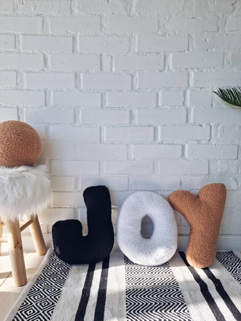 May include: Three large plush letters spelling out the word "JOY" in black, white, and brown. The letters are sitting on a black and white striped rug in front of a white brick wall.