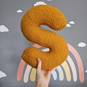 May include: A large, fuzzy, yellow letter S pillow. The pillow is being held in a hand against a gray background with a rainbow and clouds.