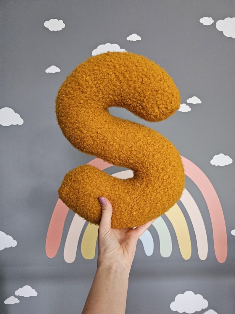 May include: A large, fuzzy, yellow letter S pillow. The pillow is being held in a hand against a gray background with a rainbow and clouds.