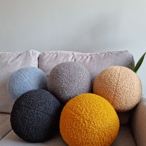 May include: Five round, fluffy pillows in different colors. The pillows are arranged in a semi-circle on a gray couch. The colors are gray, black, beige, and yellow.