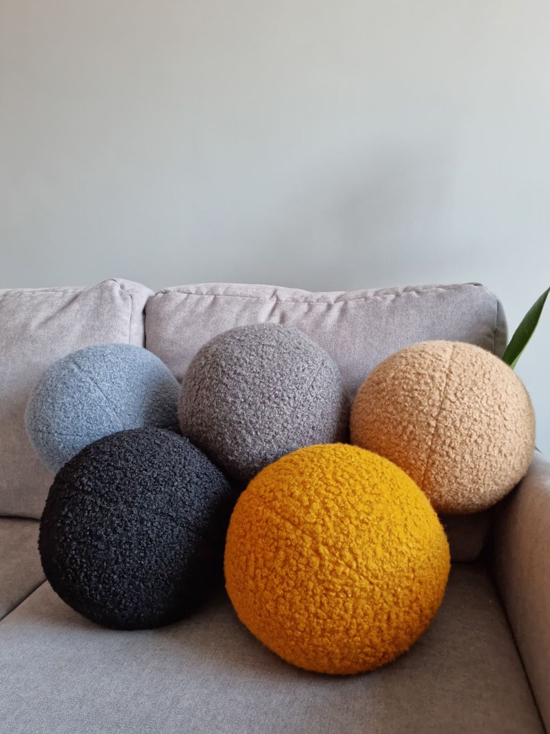 May include: Five round, fluffy pillows in different colors. The pillows are arranged in a semi-circle on a gray couch. The colors are gray, black, beige, and yellow.