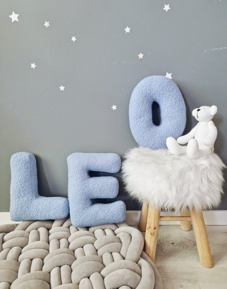 May include: A light blue plush letter O, L, and E spelling out the name "Leo" on a gray background with white stars. A white furry stool with a white teddy bear sitting on it.
