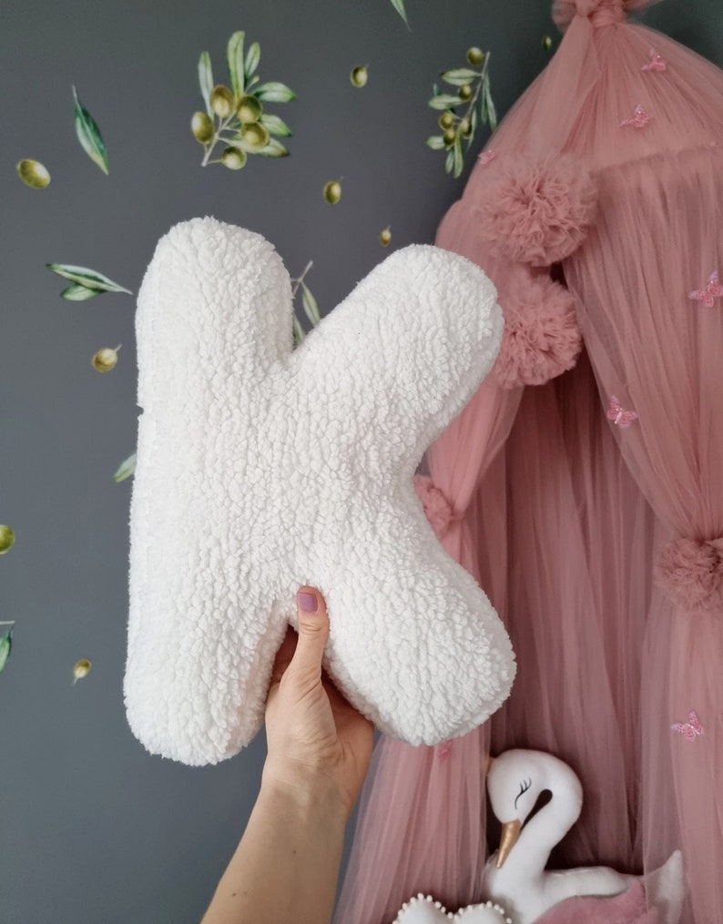 May include: White fluffy letter K pillow. The pillow is being held in a hand in front of a pink tulle canopy with white pom poms.