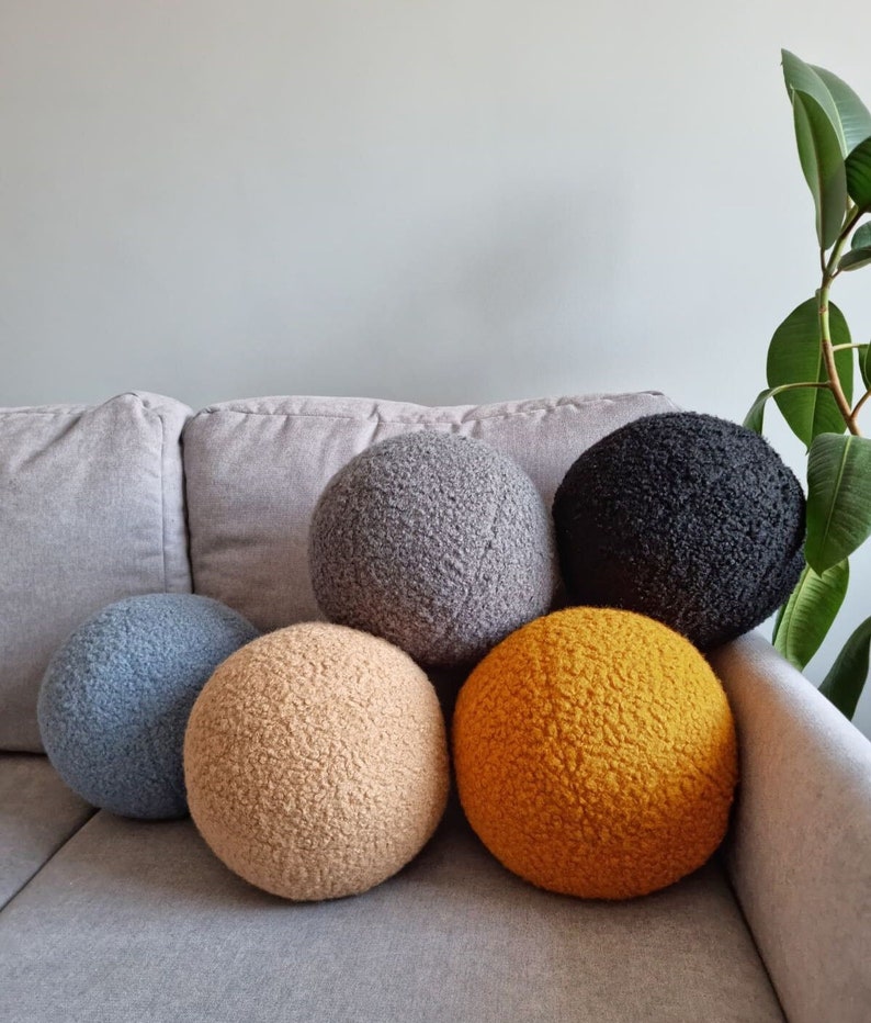 May include: Five round, fluffy, decorative pillows in various colors. The pillows are arranged in a semi-circle on a gray couch. The pillows are gray, blue, beige, and yellow.