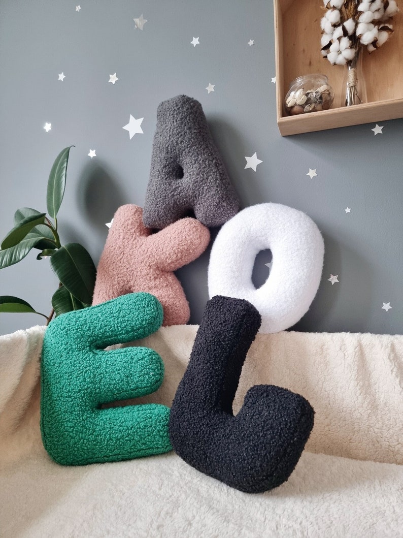 May include: Four plush letters, A, K, E, and L, in gray, pink, green, and black, spelling out the word "AKE" on a white fuzzy surface.