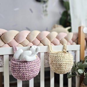 May include: Two crochet hanging baskets, one pink and one beige, attached to a white crib. The pink basket holds a white teddy bear. A braided bumper in pink and beige tones lines the crib.