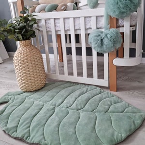 Pu&ograve; includere: Un tappetino da gioco a forma di foglia verde con una texture trapuntata, posizionato davanti a una culla bianca con biancheria da letto a motivi verdi e bianchi. La culla &egrave; decorata con un pompon di tulle verde, un cuscino a forma di nuvola verde e bianco e un cuscino bianco con un cuore verde.