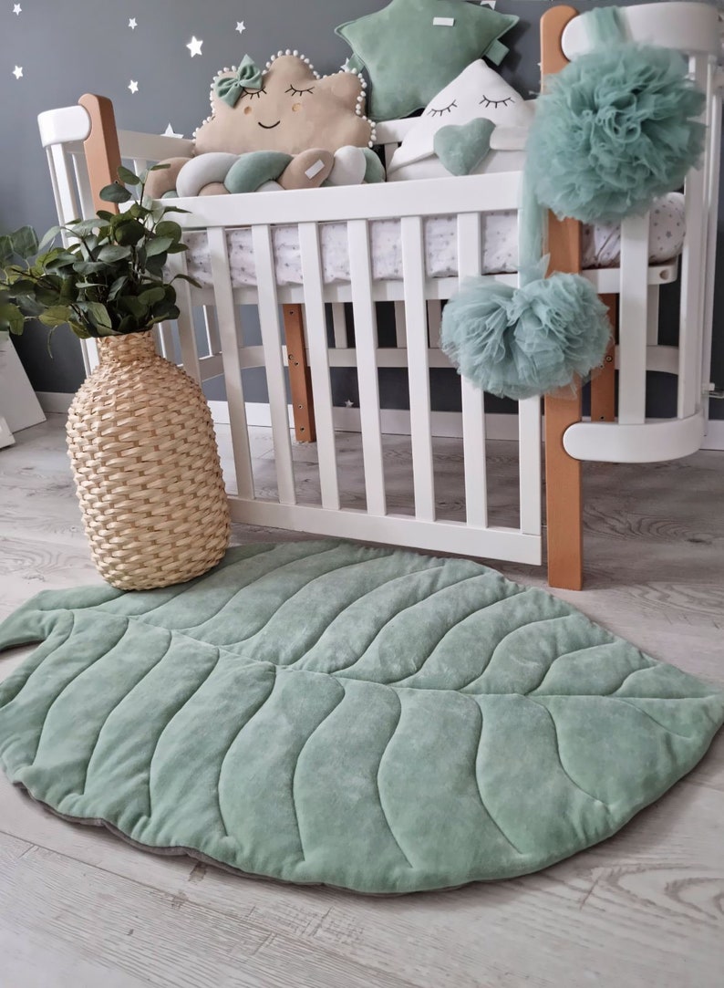 Pu&ograve; includere: Un tappetino da gioco a forma di foglia verde con una texture trapuntata, posizionato davanti a una culla bianca con biancheria da letto a motivi verdi e bianchi. La culla &egrave; decorata con un pompon di tulle verde, un cuscino a forma di nuvola verde e bianco e un cuscino bianco con un cuore verde.