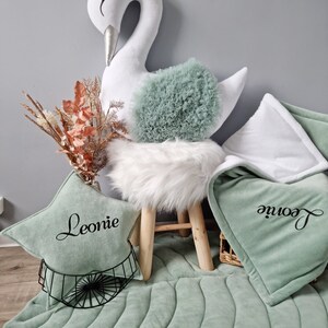 May include: A white swan plush toy with a crown on its head sits on a white faux fur stool. A green and white quilted play mat with the name "Leonie" embroidered on it is on the floor.