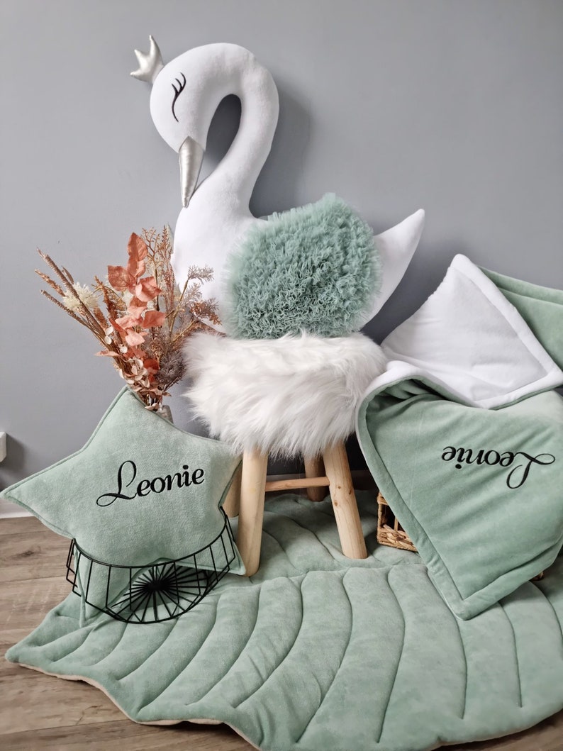 May include: A white swan plush toy with a crown on its head sits on a white faux fur stool. A green and white quilted play mat with the name "Leonie" embroidered on it is on the floor.