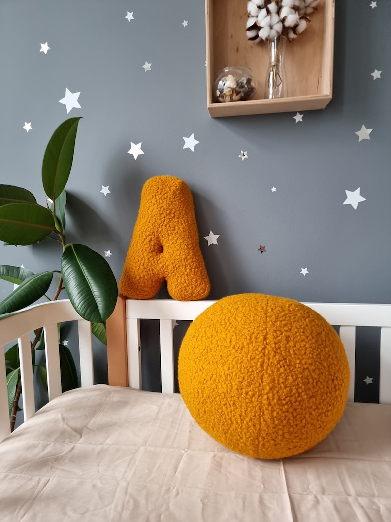 May include: A yellow plush letter A and a large yellow plush ball in a nursery setting. The wall is painted a light gray and decorated with white stars.