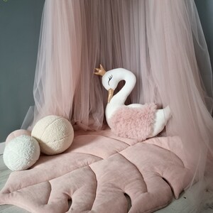 May include: A pink plush leaf-shaped play mat with a white swan plush toy and two white fluffy balls. The mat is under a pink tulle canopy.