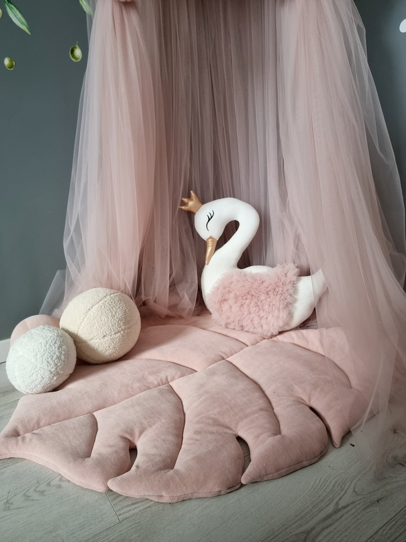 May include: A pink plush leaf-shaped play mat with a white swan plush toy and two white fluffy balls. The mat is under a pink tulle canopy.