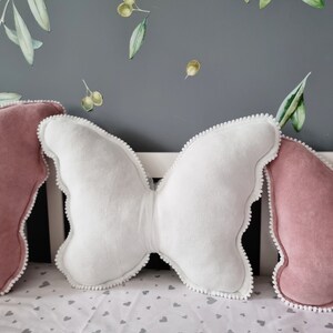 K&ouml;nnte beinhalten: Zwei dekorative Kissen, eines wei&szlig; und in Form eines Schmetterlings, und eines rosa, auf einem wei&szlig;en Bettbezug mit einem grauen Herzmuster.