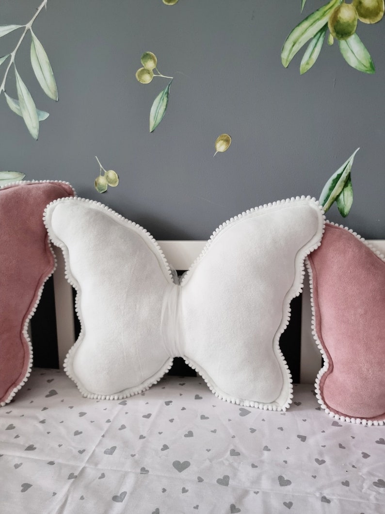 K&ouml;nnte beinhalten: Zwei dekorative Kissen, eines wei&szlig; und in Form eines Schmetterlings, und eines rosa, auf einem wei&szlig;en Bettbezug mit einem grauen Herzmuster.