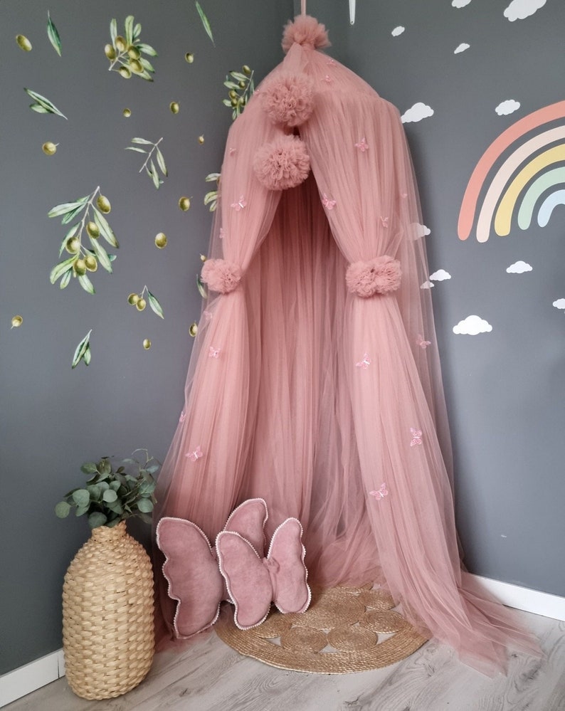 May include: A pink tulle canopy with pom poms and butterfly embellishments. The canopy is set up in a corner of a room with a rainbow wall decal and a wicker vase with greenery.