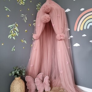 K&ouml;nnte beinhalten: Ein rosa T&uuml;ll-Baldachin mit Pompons und Schmetterlingsverzierungen. Der Baldachin h&auml;ngt von der Decke und ist von einer grauen Wand mit Olivenzweig-Aufklebern umgeben.
