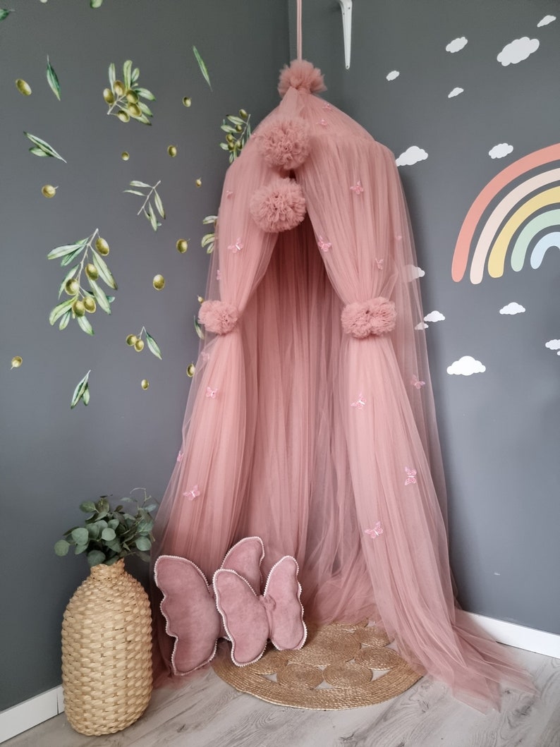 K&ouml;nnte beinhalten: Ein rosa T&uuml;ll-Baldachin mit Pompons und Schmetterlingsverzierungen. Der Baldachin h&auml;ngt von der Decke und ist von einer grauen Wand mit Olivenzweig-Aufklebern umgeben.