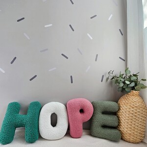 May include: The word "HOPE" spelled out in large, plush letters. The letters are green, white, pink, and green. The letters are sitting on a white surface in front of a light green wall with gray and white confetti-like shapes.