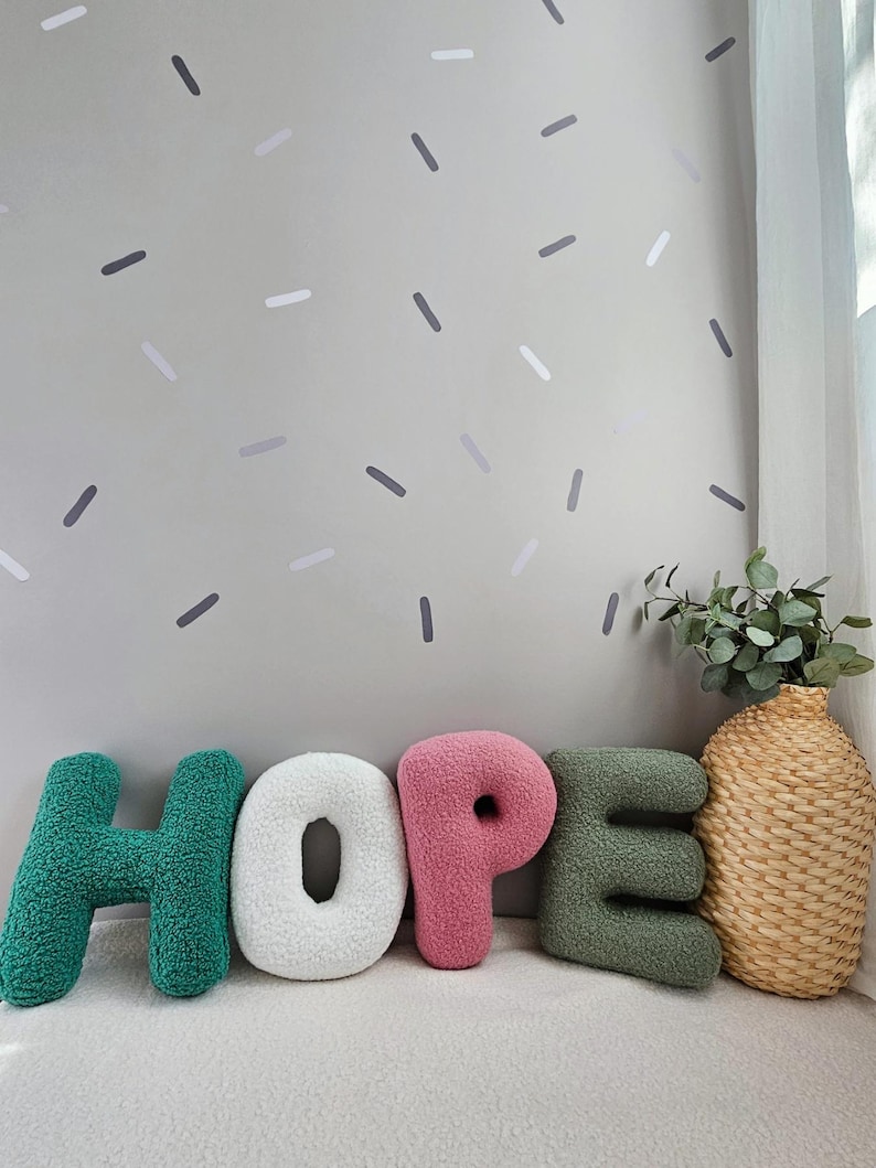 May include: The word "HOPE" spelled out in large, plush letters. The letters are green, white, pink, and green. The letters are sitting on a white surface in front of a light green wall with gray and white confetti-like shapes.