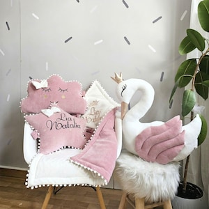 Puede incluir: Una colección de decoración de guardería rosa y blanca, que incluye almohadas en forma de nube y estrella, un peluche de cisne con corona y una manta. Las almohadas tienen detalles bordados y el nombre "Lia Natalia". El cisne tiene alas rosas.