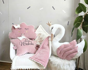 May include: A collection of pink and white nursery decor, including cloud and star-shaped pillows, a swan plush toy with a crown, and a blanket. The pillows have embroidered details and the name "Lia Natalia". The swan has pink wings.