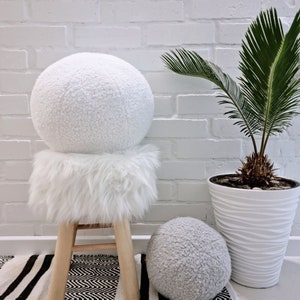 May include: Two white fluffy round poufs, one on a wooden stool with a white fluffy seat cushion, and the other on the floor. The stool and poufs are in front of a white brick wall with a potted palm plant.