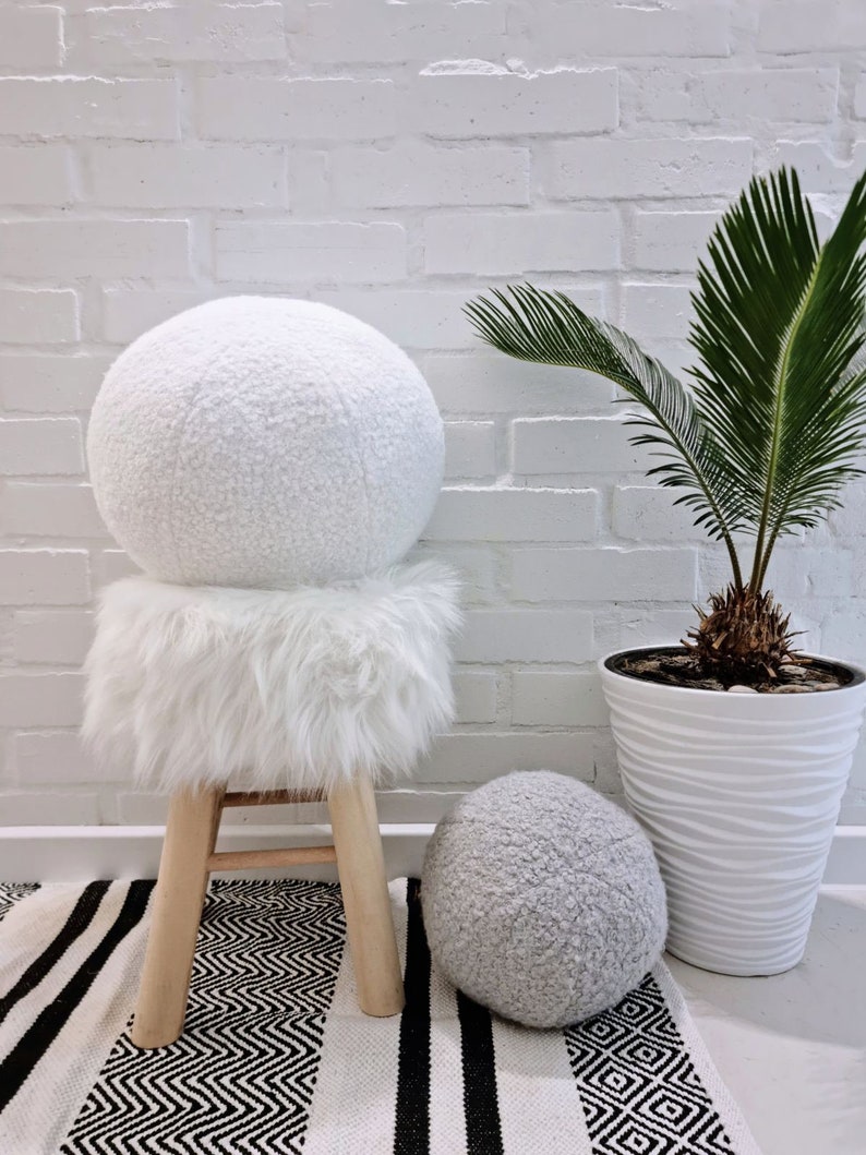 May include: Two white fluffy round poufs, one on a wooden stool with a white fluffy seat cushion, and the other on the floor. The stool and poufs are in front of a white brick wall with a potted palm plant.