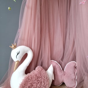 May include: A white swan-shaped pillow with a pink tulle ruffle and a gold crown. The pillow is sitting on a brown woven rug in front of a pink tulle curtain.