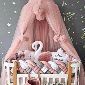 Peut inclure: Un lit bébé blanc avec un pare-chocs tressé rose et gris, une canopée en tulle rose avec des pompons, un oreiller rose et blanc avec le texte "Little Princess", un jouet en peluche de cygne blanc et un panier en tricot rose.