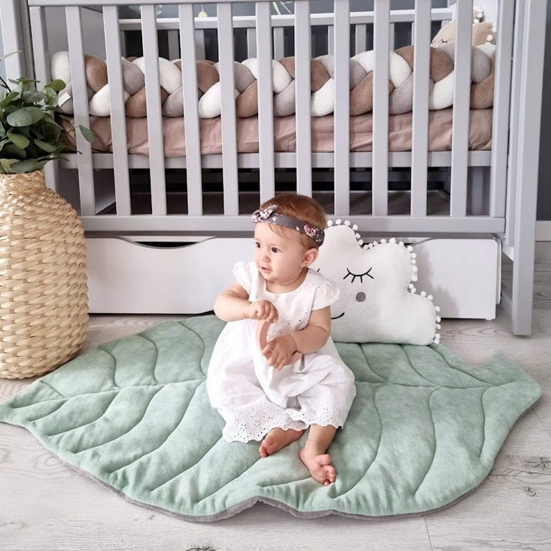 Pu&ograve; includere: Una bambina con un vestito bianco e una fascia floreale &egrave; seduta su un tappetino da gioco verde a forma di foglia. Il tappetino ha una texture trapuntata e un bordo grigio. Un cuscino bianco a forma di nuvola con un disegno di occhio nero &egrave; dietro la bambina.