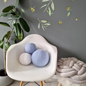 May include: Three soft, round pillows in a light blue and white color scheme. The pillows are sitting on a white chair with wooden legs. A gray braided pouf sits on a wooden stool with a white faux fur seat.