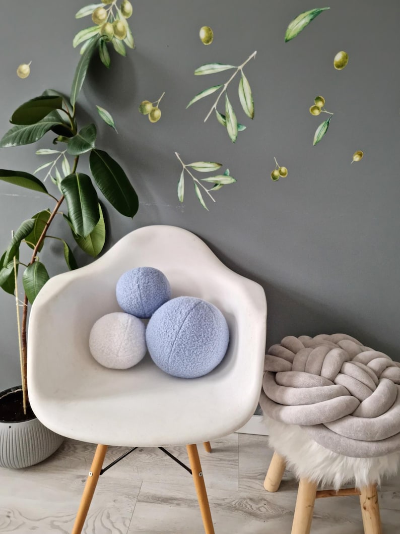 May include: Three soft, round pillows in a light blue and white color scheme. The pillows are sitting on a white chair with wooden legs. A gray braided pouf sits on a wooden stool with a white faux fur seat.