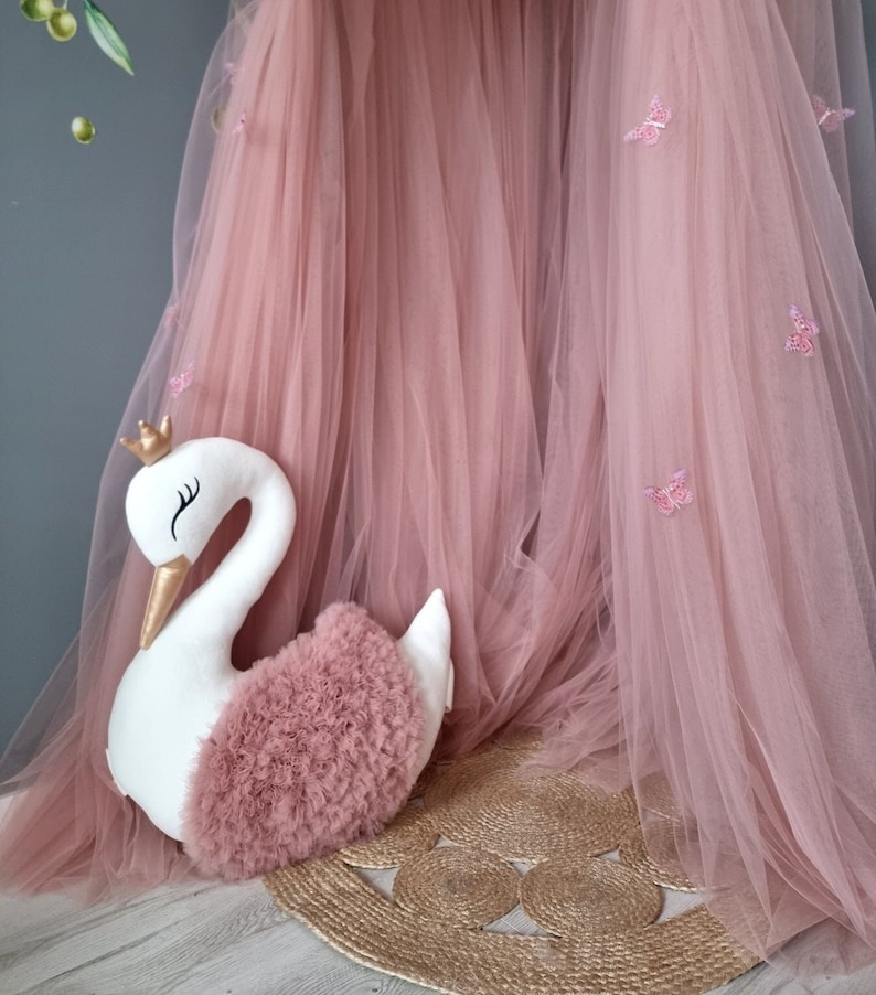 May include: A white swan-shaped pillow with a pink tulle ruffle and a gold crown. The pillow is sitting on a woven rug in front of a pink tulle canopy.
