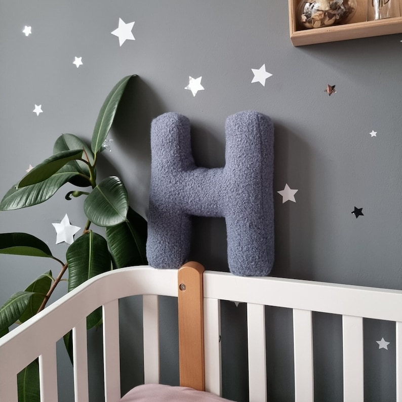 May include: A light blue plush letter H pillow against a gray wall with white stars. The pillow is sitting on a white crib with a wooden side rail.