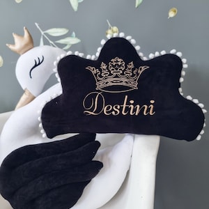 May include: White and black swan plush toy with a black pillow that says "Destini" in gold embroidery with a gold crown above the name. The pillow has white pom-poms around the edge.