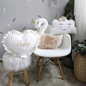 May include: A selection of nursery cushions. The cushions include a white heart-shaped pillow embroidered with "Baby" in gold, a swan with a gold crown, a cloud with a smiling face, and a fluffy beige cushion. They are arranged on a white chair and a small stool.