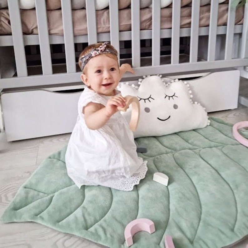 Pu&ograve; includere: Una bambina con un vestito bianco e una fascia floreale &egrave; seduta su un tappetino da gioco verde a forma di foglia. Dietro di lei c'&egrave; un cuscino bianco a forma di nuvola con un viso sorridente e ciglia.
