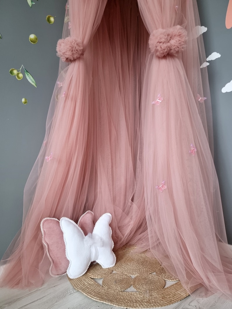 K&ouml;nnte beinhalten: Ein rosa T&uuml;ll-Baldachin mit Schmetterlingsverzierungen und zwei Bommeln oben. Zwei schmetterlingsf&ouml;rmige Kissen liegen auf dem Boden vor dem Baldachin.