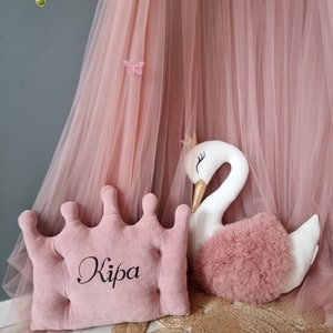 May include: A pink crown-shaped pillow with the name "Kipa" embroidered on it. The pillow is next to a white swan plush toy with a pink tulle skirt. The background is a pink tulle canopy.