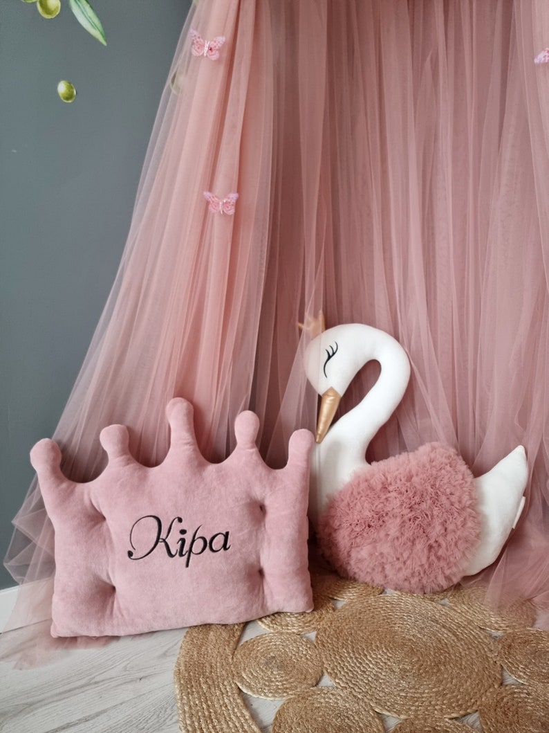 May include: A pink crown-shaped pillow with the name "Kipa" embroidered on it. The pillow is next to a white swan plush toy with a pink tulle skirt. The background is a pink tulle canopy.
