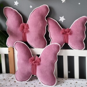 May include: Three pink butterfly-shaped pillows with white trim and a tulle bow on each. The pillows are arranged on a white crib.