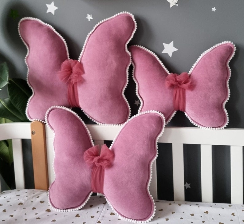 May include: Three pink butterfly-shaped pillows with white trim and a tulle bow on each. The pillows are arranged on a white crib.