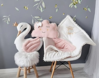 May include: A nursery scene featuring a white swan-shaped pillow with a gold crown, a pink cloud-shaped pillow, and a white chair with a pink cushion. The chair has wooden legs. A small, white, furry stool and a round rug are also present.