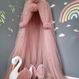 May include: A pink tulle canopy with pom poms and butterfly accents. A white swan plush toy and a pink butterfly pillow are in front of the canopy.