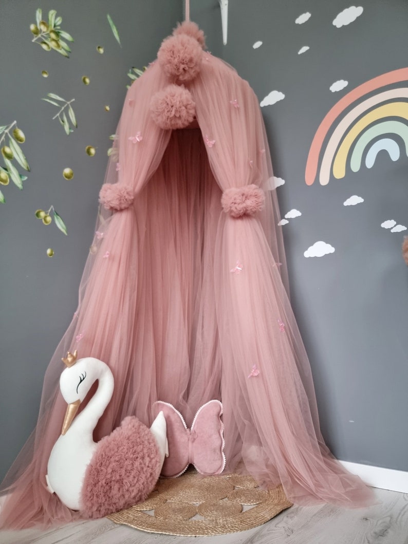 May include: A pink tulle canopy with pom poms and butterfly accents. A white swan plush toy and a pink butterfly pillow are in front of the canopy.