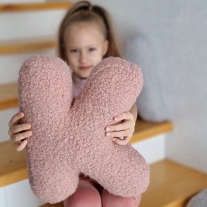 May include: A pink fluffy letter K pillow being held by a child.